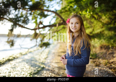 Cute little girl standing by the lake enjoying beautiful sunset view. Child exploring nature. Family activities in summer. Stock Photo