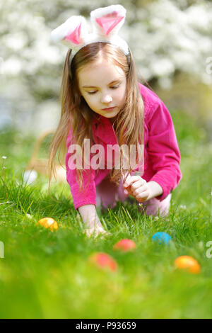 Adorable little girl hunting for easter egg in blooming spring garden on Easter day. Cute child celebrating Easter outdoors. Stock Photo