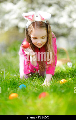 Adorable little girl hunting for easter egg in blooming spring garden on Easter day. Cute child celebrating Easter outdoors. Stock Photo