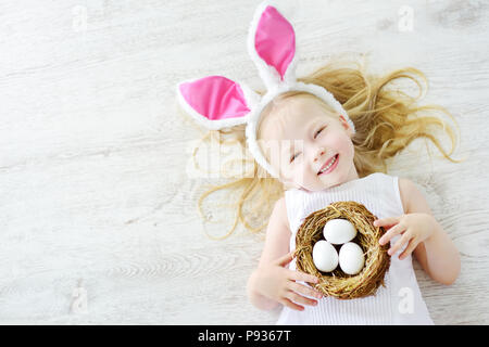 Cute little girl wearing bunny ears playing egg hunt on Easter. Adorable child celebrate Easter at home. Stock Photo