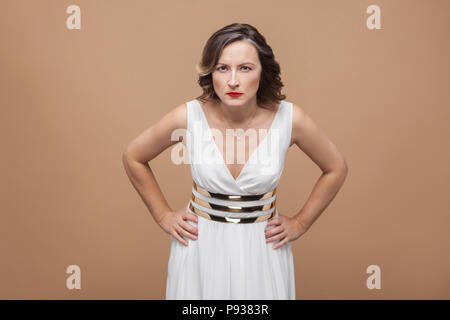 Closeup portrait of angry woman looking at camera. Emotional expressing woman in white dress, red lips and dark curly hairstyle. Studio shot, indoor,  Stock Photo