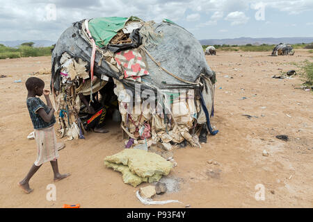 Kakuma, Kenya - On the edge of the refugee camp Kakuma. A girl is standing in front of a hut covered with old plastic tarpaulins, blankets, boxes and garbage. Stock Photo