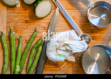 Cucumbers, green beans, measuring utensils and a knife lie on a wooden cutting board during food preparation. Stock Photo