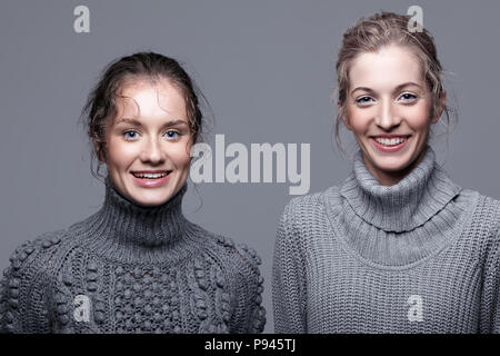 Two young happy smiling women in gray sweaters on grey studio background. Beautiful girls stretching hands forward in embrace. Female friendship conce Stock Photo