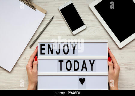 Workplace with tablet, smartphone, notepad and 'Enjoy today' word on lightbox over white wooden background, top view. Female hands hold lightbox. From Stock Photo