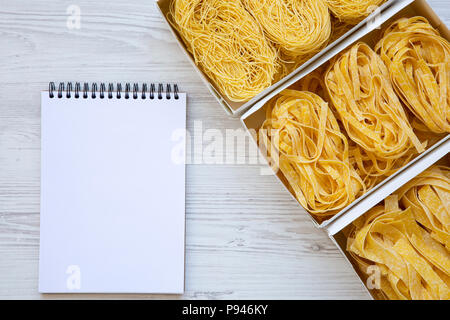 Various uncooked pasta in paper boxes with notebook on white wooden table, top view. From above. Stock Photo