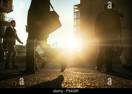 People walking on city street with sun flare in the background. Men commuting to office early in the morning carrying office bags. Stock Photo