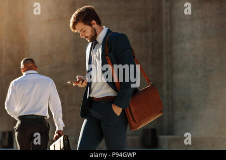 Men in formal clothes commuting to office early in the morning carrying office bags. Businessman using mobile phone while walking on city street to of Stock Photo