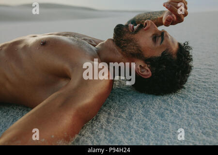 Tired man lying on sand dune after intense physical training. Athlete resting on sand after workout session. Stock Photo