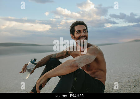 Smiling muscular male athlete sitting on desert sand with a water ball in hand. Tired man taking a break from training in desert. Stock Photo