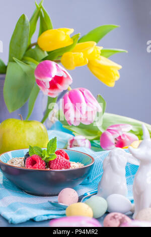 Delicious spring breakfast on a background of gray stone. Bouquet of fresh tulips. Small and large colored Easter eggs. Oatmeal, coffee, fresh berries. Postcard for Easter. Stock Photo