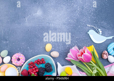 Delicious spring breakfast on a gray stone background. A bouquet of fresh tulips of pink and mint color. Small and large colored easter eggs. Oatmeal, biscuits, coffee, fresh raspberries and blackberr. Stock Photo