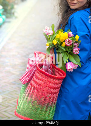 girl with a bouquet of spring tulips in pink. In a blue raincoat and yellow boots on the street in Europe. Happy woman. Smile on the face. Card. Stock Photo