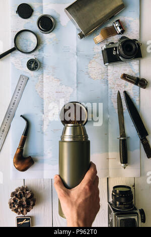 Adventure planning flat lay. Travel vintage gear on map. Traveler, explorer hands in frame pouring coffee or tea from thermos into mug. Vertical explo Stock Photo