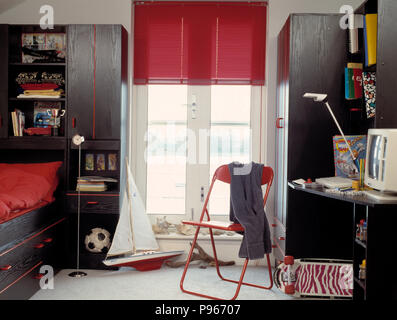 Model sailing boat and red metal chair in front of French windows with red blind in child's bedroom with black units Stock Photo