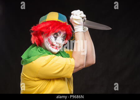 Scary clown with a horrible make-up laughs and with big knives in hands on a black background Stock Photo