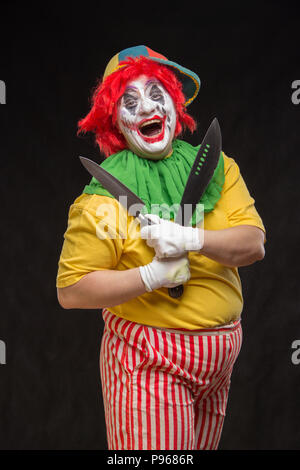 Scary clown with a horrible make-up laughs and with big knives in hands on a black background Stock Photo