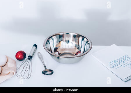 Kitchen accessories and products for baking on a white table: a metal bowl, a whisk and a spoon next to a recipe, an egg and ripe plum. Stock Photo