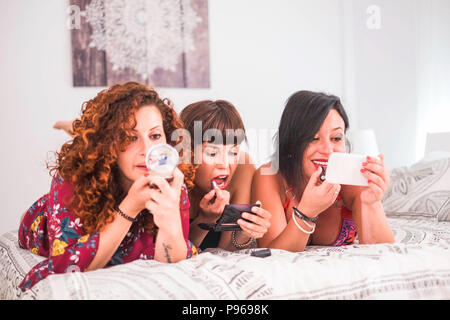 three women friends doing make up at home in friendship lay down on the bed in the bedroom. making pretty and being beauty for young caucasian females Stock Photo