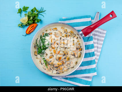 Mussels in milk cream y sauce with aromatic herbs and lemon in red cooking pan. Blue background Stock Photo