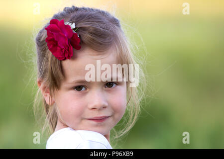 Portrait of beautiful small preschool blond girl with nice gray eyes and red rose in hair smiling dreamily in camera on blurred bright green backgroun Stock Photo