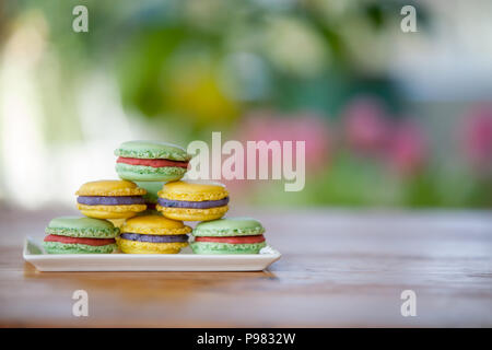 Macarons on a plate on a wooden table with blurry flowers and plants in the background Stock Photo