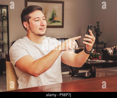 Man pointing the finger to the smartphone. Man with happy expression looking to the phone. At home, wearing casual clothes. Stock Photo