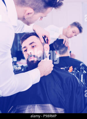 Pleasant  male hairdresser accurately cutting beard of client at hair salon Stock Photo