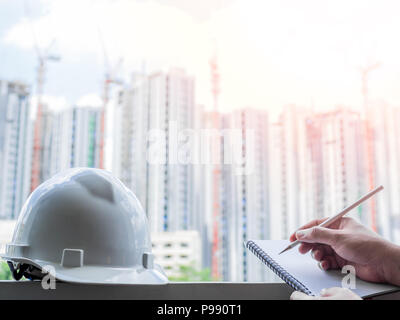 White hard safety helmet for civil engineer, protect and safety hat for engineering worker and hand writing notebook by pencil on construction buildin Stock Photo