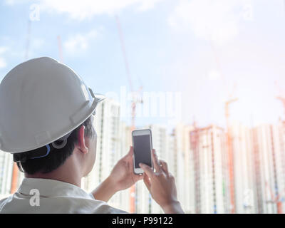 Engineer man back view wearing white hard safety helmet and white shirt capture a photo by mobile phone on construction building background on sunshin Stock Photo