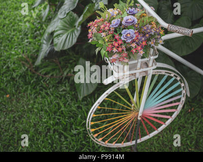 White vintage bicycle with colourful wheel and colourful flowers in the white pot in the basket on green grass with tropical palm leaves in the garden Stock Photo