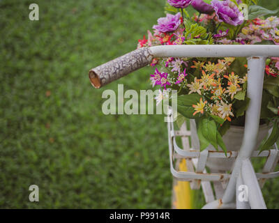 White vintage bicycle and colourful flowers in the white pot in the basket on green grass in the garden. Decoration in park with space. Stock Photo