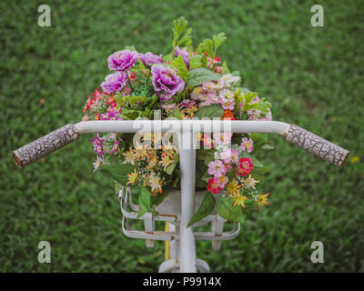 White vintage bicycle and colourful flowers in the white pot in the basket on green grass in the garden. Decoration in park. Stock Photo