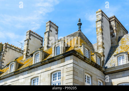 The top of a residential building in the old city of Saint-Malo with a slate roof covered with lichen, dormer windows and chimneys against blue sky. Stock Photo