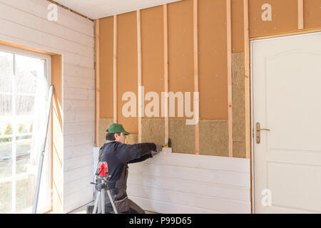 The worker makes finishing works of walls with a white wooden board, using laser line level. Building heat-insulating eco-wooden frame house with wood fiber plates Stock Photo