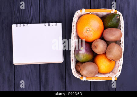 Assorted fruits in basket, top view. Fresh tropical fruits in wicker basket and blank spiral notebook on dark wooden background. Diet plan concept. Stock Photo