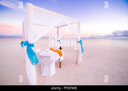 Romantic dinner table set-up for a honeymoon couple on the beach under sunset sky. Exotic wedding and honeymoon concept Stock Photo