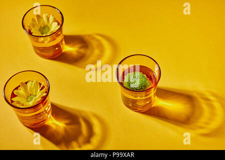 Yellow transparent glasses with water and yellow and green flowers on a zolot paper background with a pattern from the shadows. Top view Stock Photo