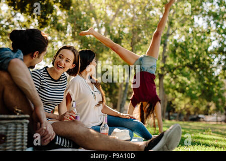 Group of friends sitting at park with a woman doing a cartwheel jump in background. Young man and women enjoying a summer day at park. Friends enjoyin Stock Photo