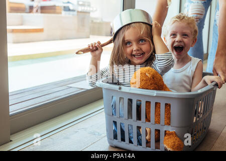 Excited children sitting in a washing basket being pushed by mother. Girl wearing a bowl as helmet with boy laughing. Stock Photo