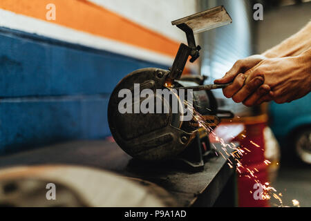 Hands of mechanic grinding a vehicle part on bench grinder in workshop. Auto mechanic using electrical grinding machine in service station. Stock Photo