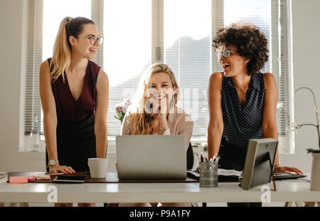 Smiling young women at work desk at office. Group of three diverse women in office smiling. Stock Photo
