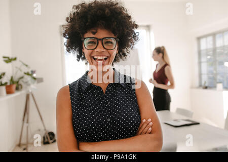 Portrait of young african businesswoman standing with her arms crossed in meeting room with colleague giving presentation in background. Woman laughin