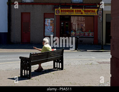 Senior woman sitting on a park bench, smoking a cigarette, Dalton, Cumbria, England UK Stock Photo