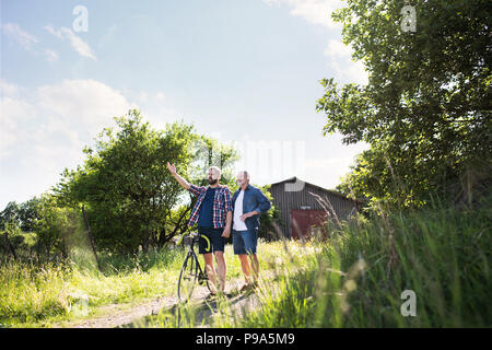 An adult hipster son with bicycle and senior father walking in sunny nature. Stock Photo