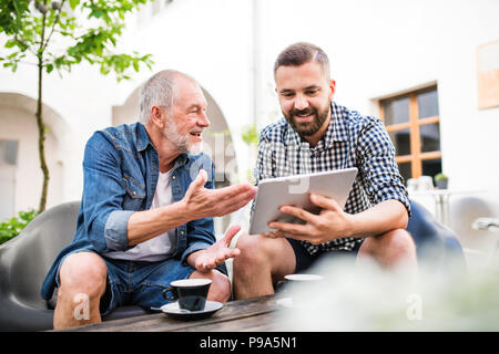 An adult hipster son with tablet and senior father in a cafe in town. Stock Photo