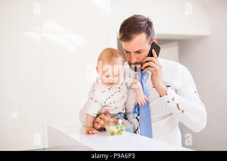 Father with shirt and tie and smartphone feeding a baby son at home, when talking on the phone. Stock Photo