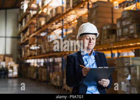 Senior woman warehouse manager or supervisor with white helmet and clipboard. Stock Photo