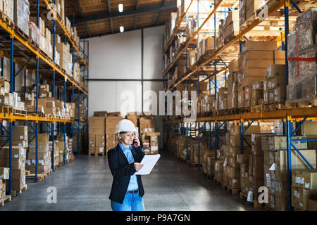 Senior woman warehouse manager or supervisor with tablet and smartphone, making a phone call. Stock Photo