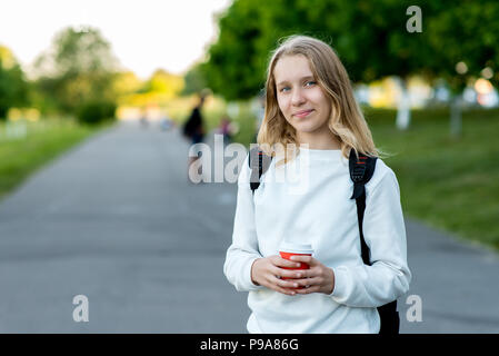 Teenage girl in summer outdoors. In his hands holds cup of coffee. Behind his backpack. Free space for text. Lunch break, rest after school. Stock Photo